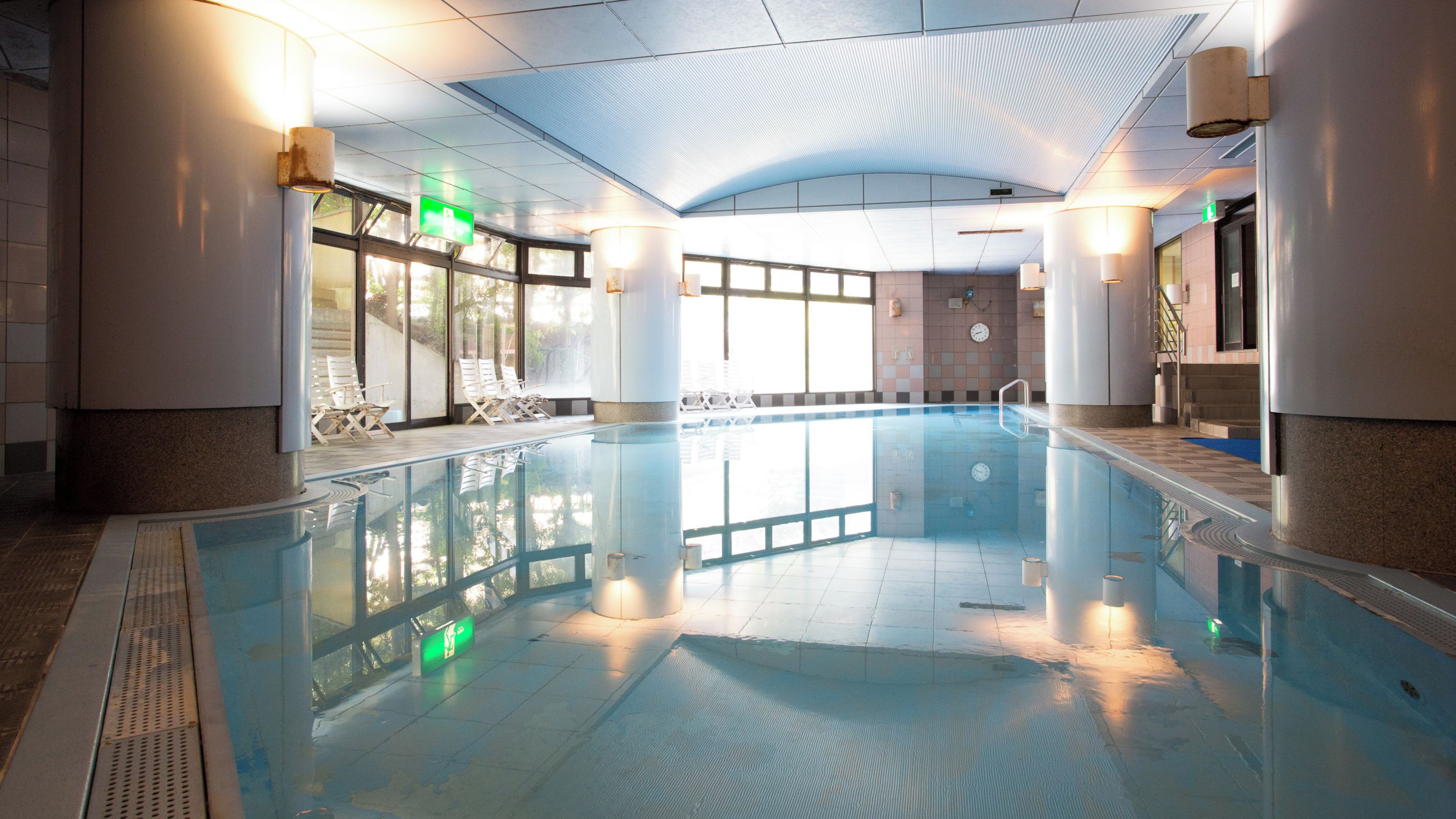 An indoor swimming pool with clear glass walls, a tiled floor, and a ceiling with a ribbed design.