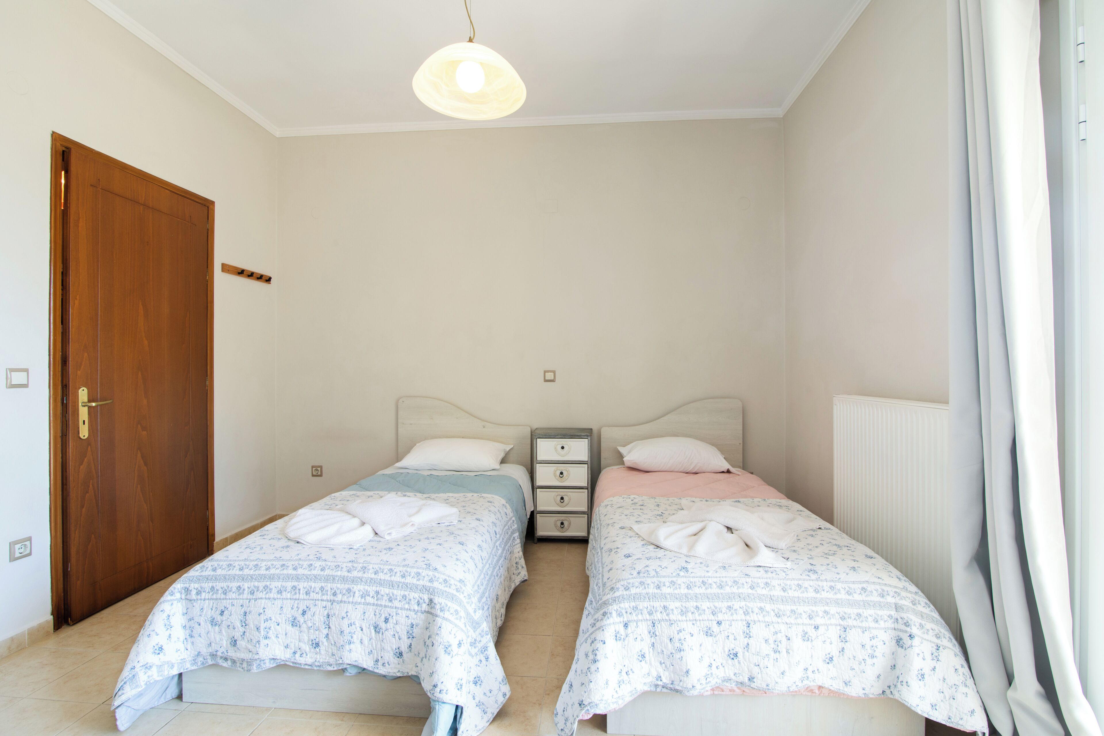 A bedroom with two single beds, a wooden door, a bedside table with drawers, and a hanging light fixture.