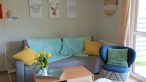 Ein Wohnzimmer mit grauem Sofa, blauem Sessel, einem Holztisch und einem kleinen weißen Hocker. An den Wänden hängen drei Kunstwerke und es steht eine Stehlampe.