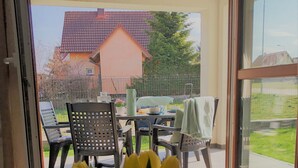 Blick durch eine Glastür auf einen Außenbereich mit Terrasse, inklusive Tisch, Stühlen, einem weißen, flauschigen Teppich und einem Strauß gelber Tulpen.