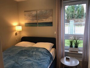 Une chambre à coucher comprenant un lit, une table de chevet, une lampe fixée au mur, une fenêtre avec des rideaux et une vue sur la verdure à l’extérieur.