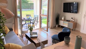 Ein modernes Wohnzimmer mit Sofa, Couchtisch und Sesseln, ein Essbereich mit Tisch und Stühlen, ein an der Wand befestigter Fernseher und Blick ins Freie durch große Glasüren.