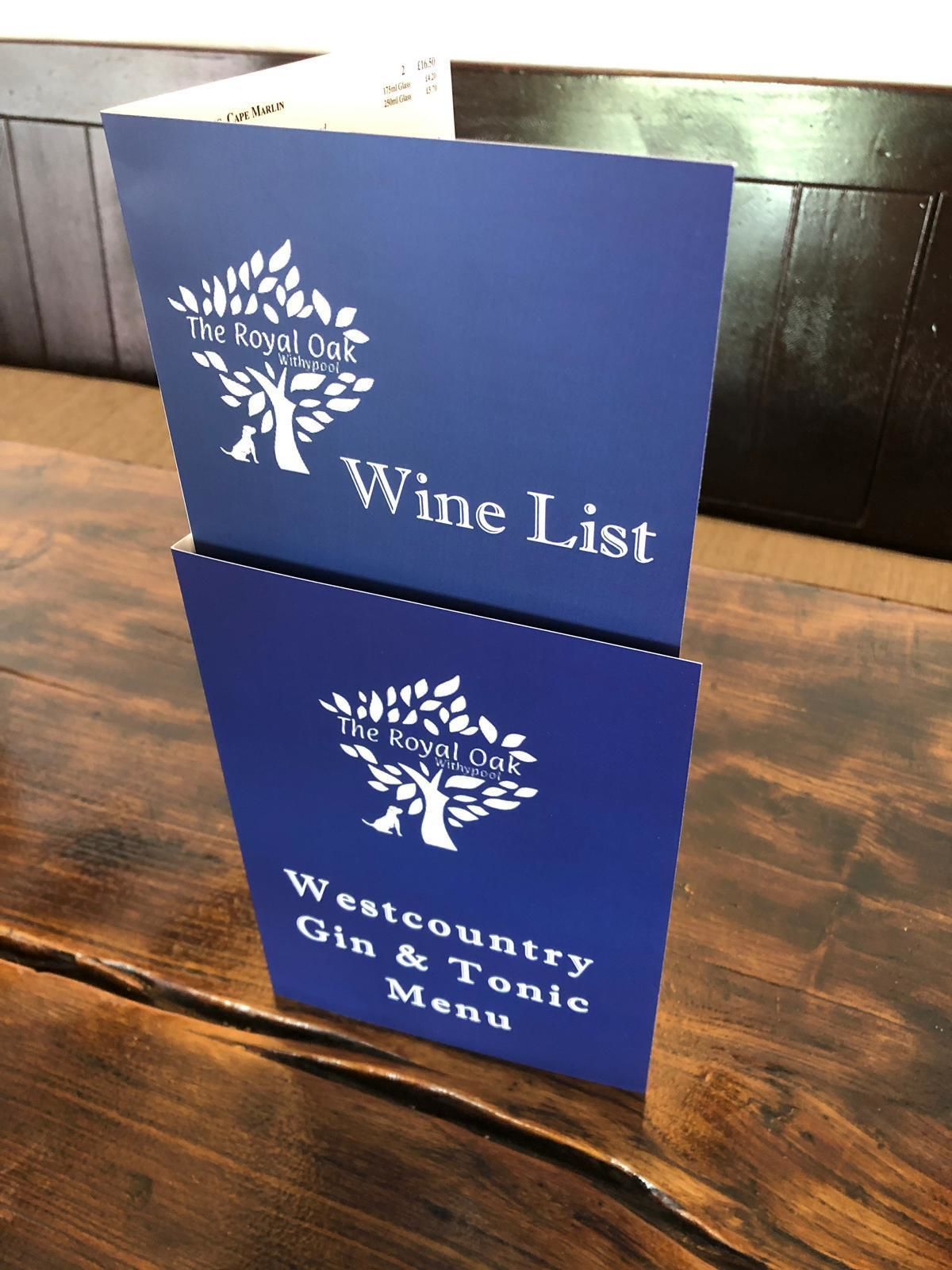 Two blue signs stacked on a wooden table, one labeled "The Royal Oak Wine List" and the other "Westcountry Gin & Tonic Menu".