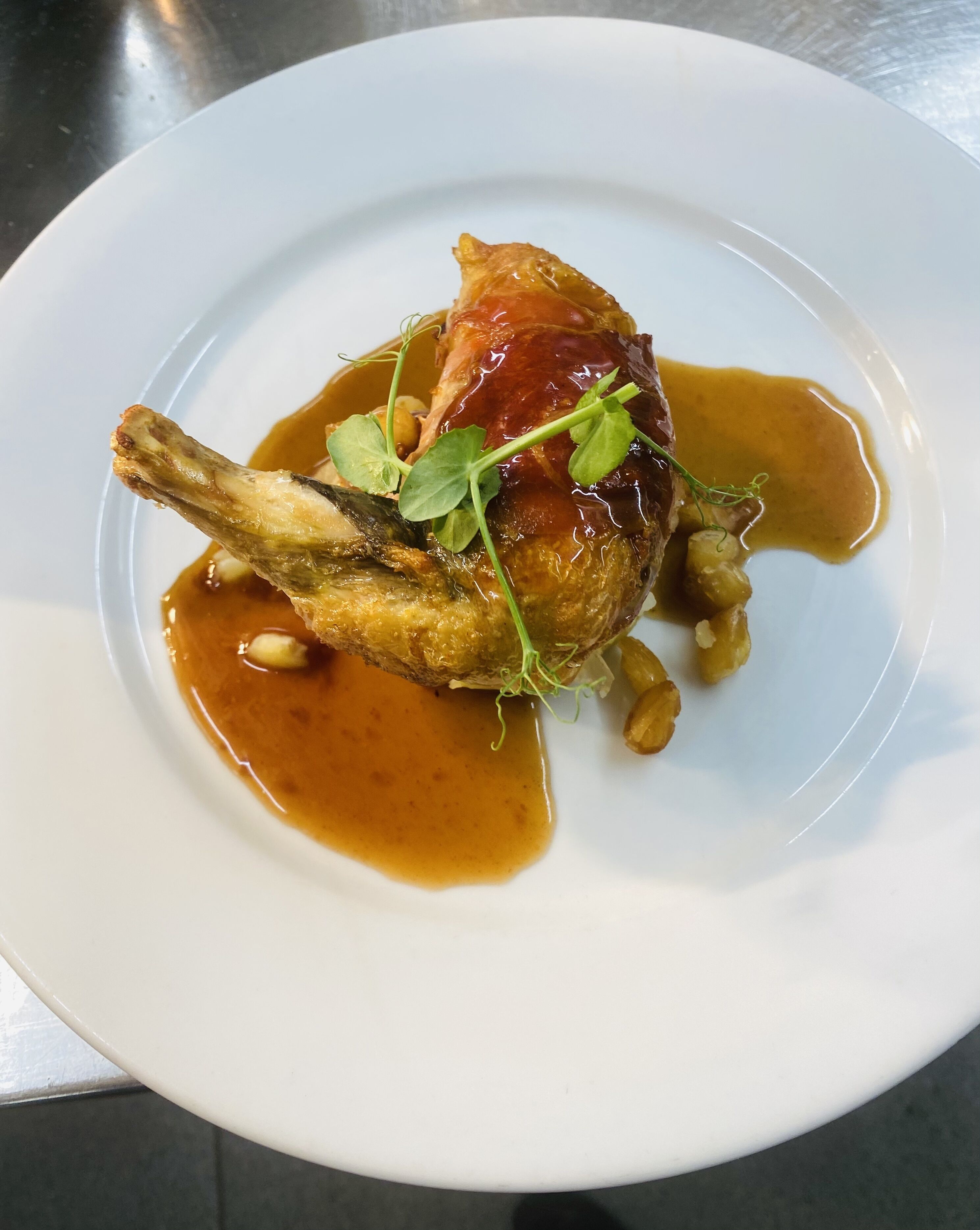 A white plate with a roasted duck leg, garnished with greens and sauce.