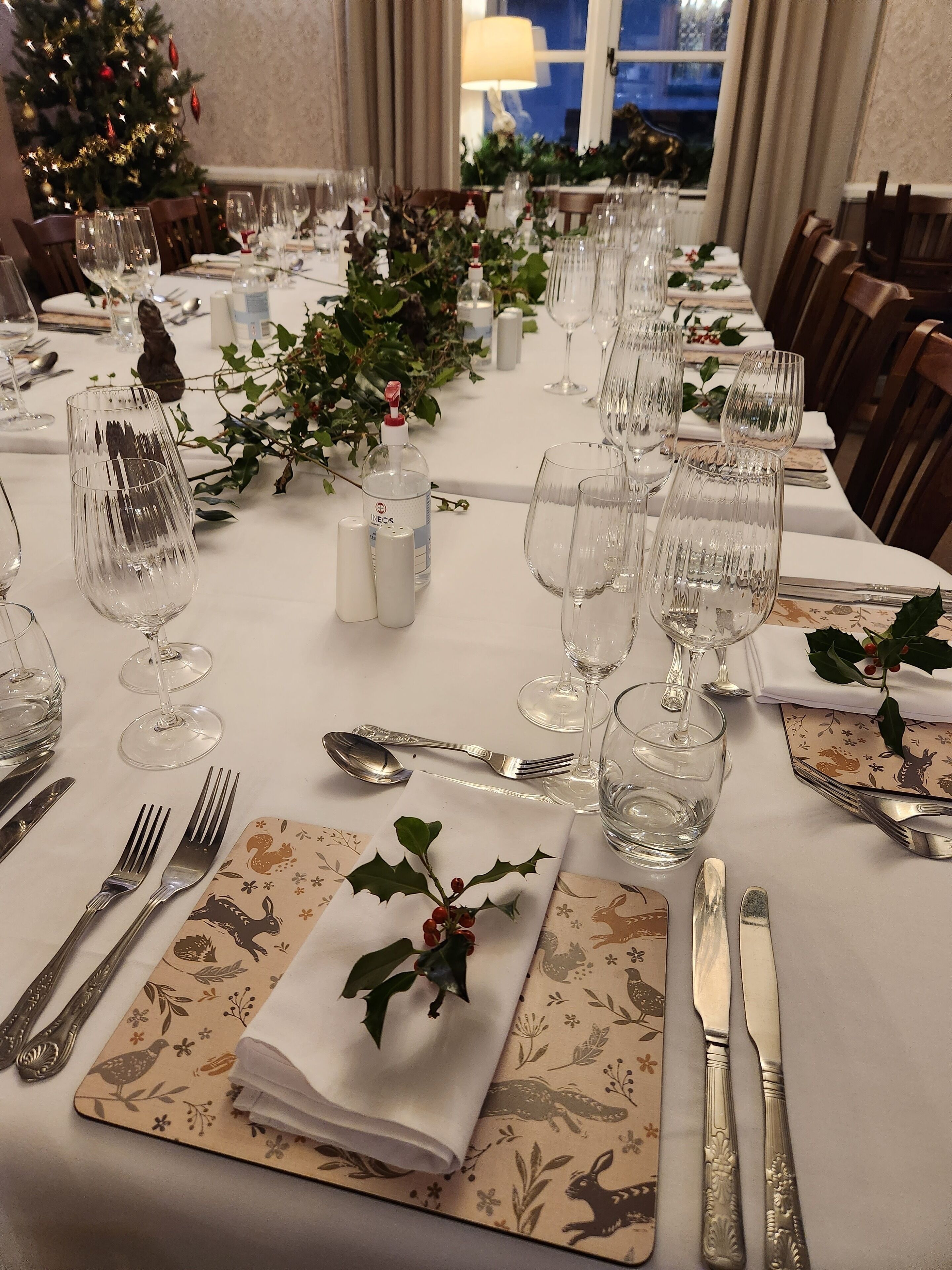 Mesa de comedor dispuesta con varias copas de vino, cubiertos y elementos decorativos como acebo y un florero.