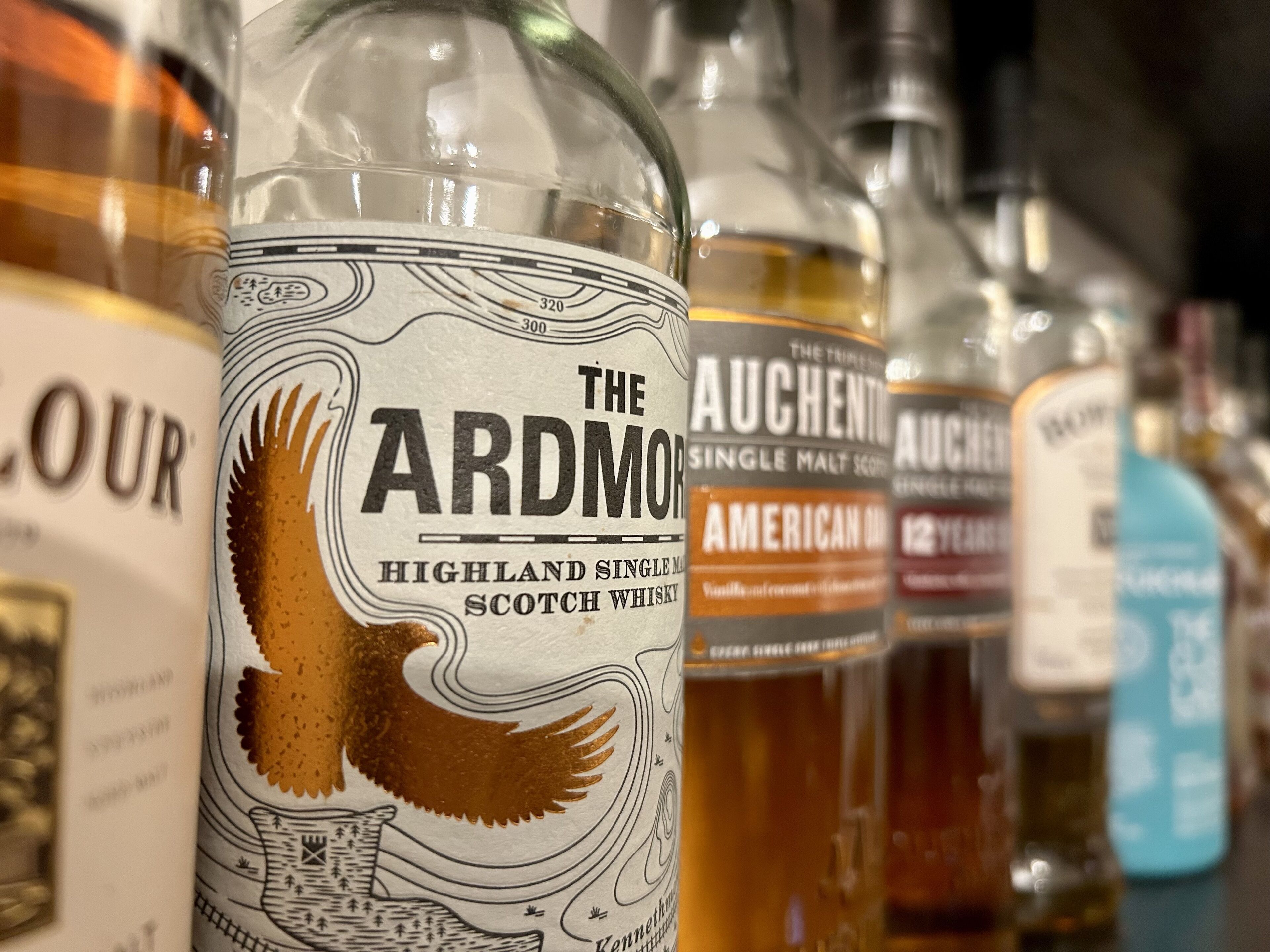 A row of whisky bottles with labels, including 'The Ardmo Highland Single Malt Scotch Whisky'.