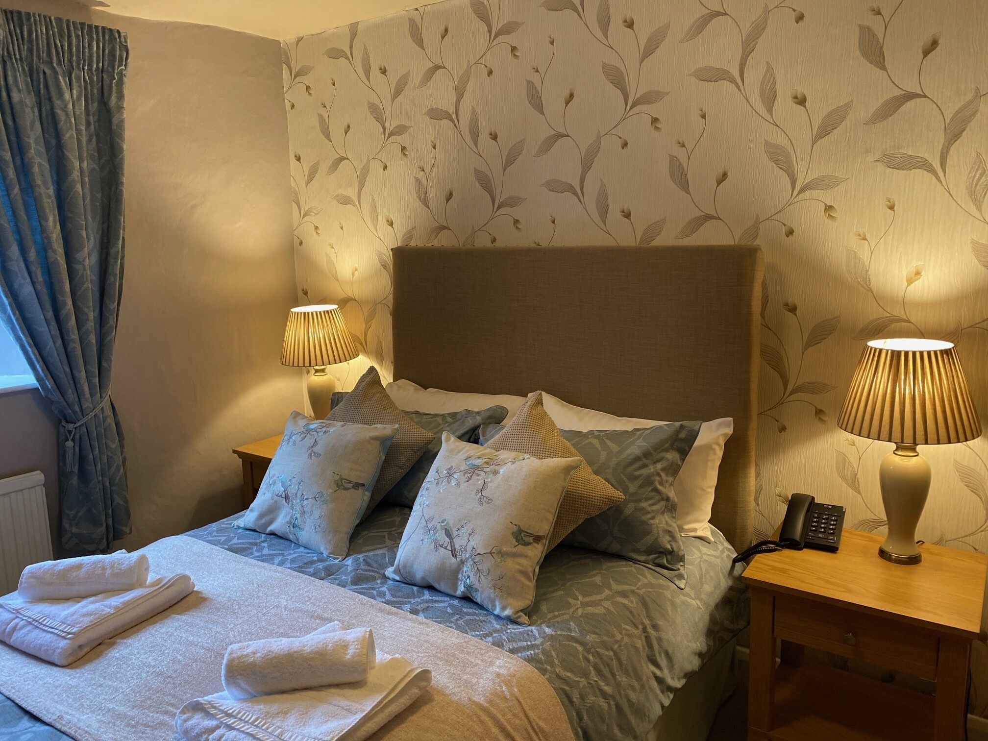 Una cama bien hecha con almohadas estampadas, una lámpara de noche y un teléfono sobre una mesita de madera.