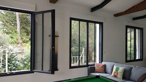 Una mesa de billar en una habitación con ventanas amplias, un sofá gris con cojines coloridos y vistas a árboles y un balcón.