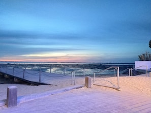 Une promenade en bois qui mène à une plage, d’où l’on peut admirer l’océan et apercevoir un pont au loin.