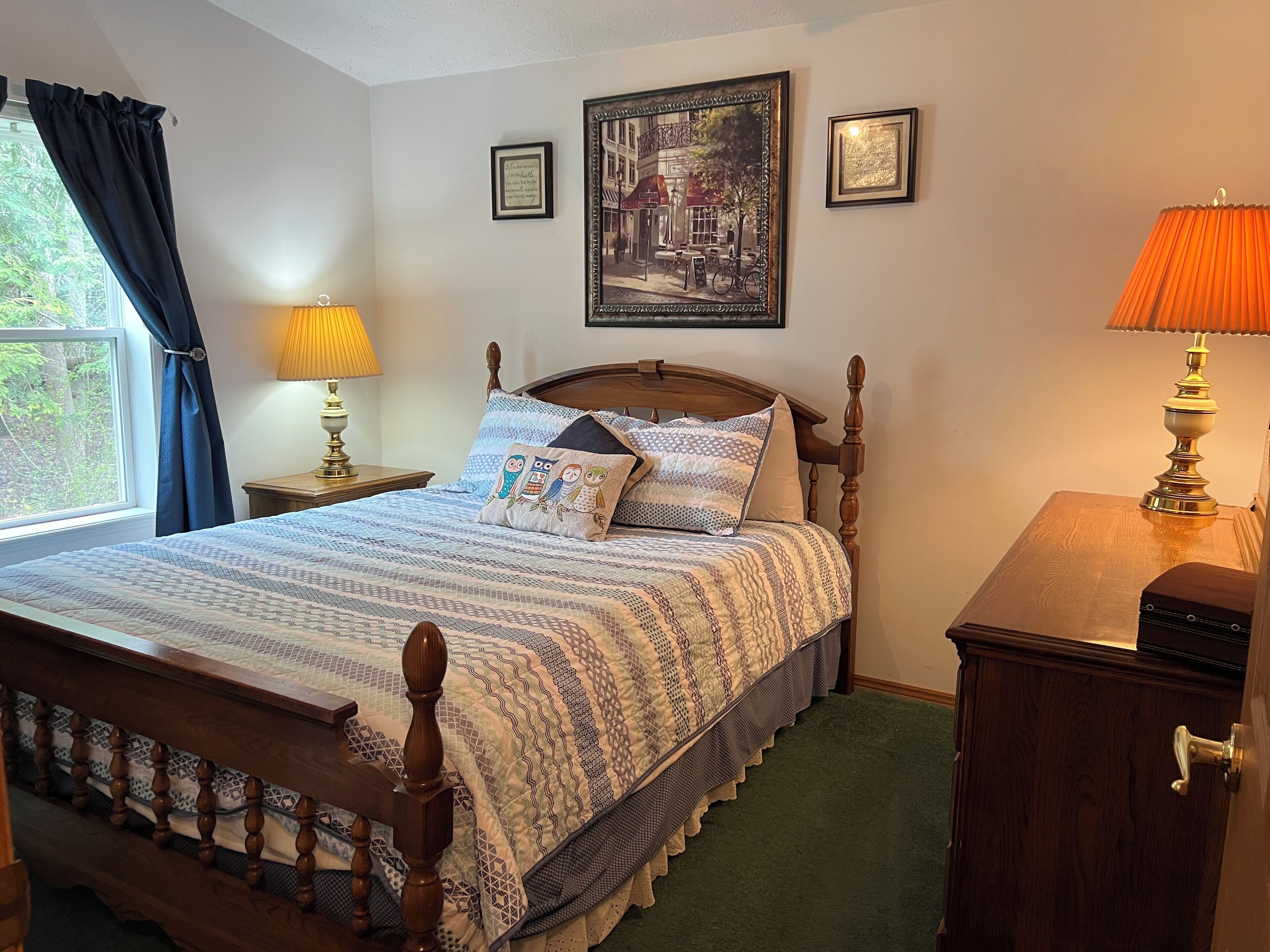 Un dormitorio ordenado con una cama de madera, dos lámparas de noche, una ventana con cortinas y un cuadro en la pared.