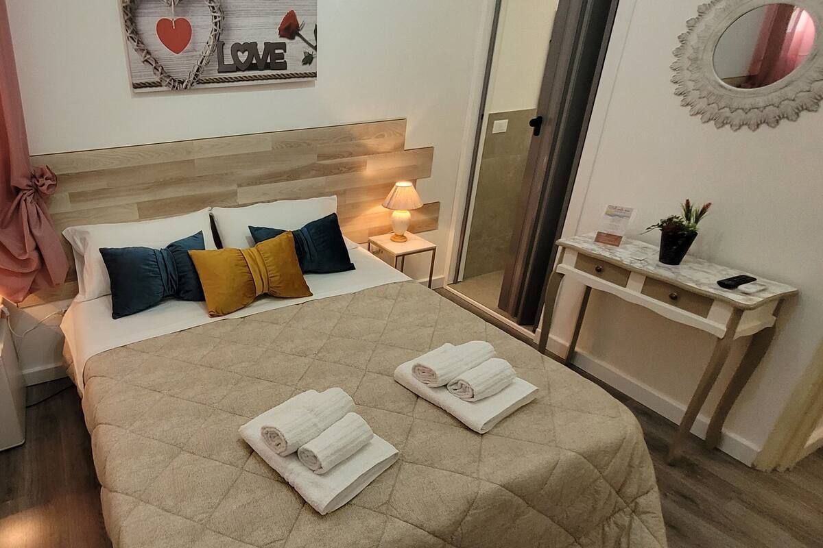 Una habitación de hotel con una cama, lámparas de noche, una mesita con una planta y un espejo en la pared.