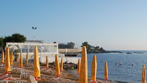 Une plage bordée de rangées de parasols et de chaises longues, un bâtiment à la façade en verre, et des bateaux sur l’eau.