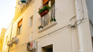 Une rue étroite bordée d’un immeuble doté d’un balcon orné de plantes en pot, de climatiseurs et d’une porte avec un paillasson.