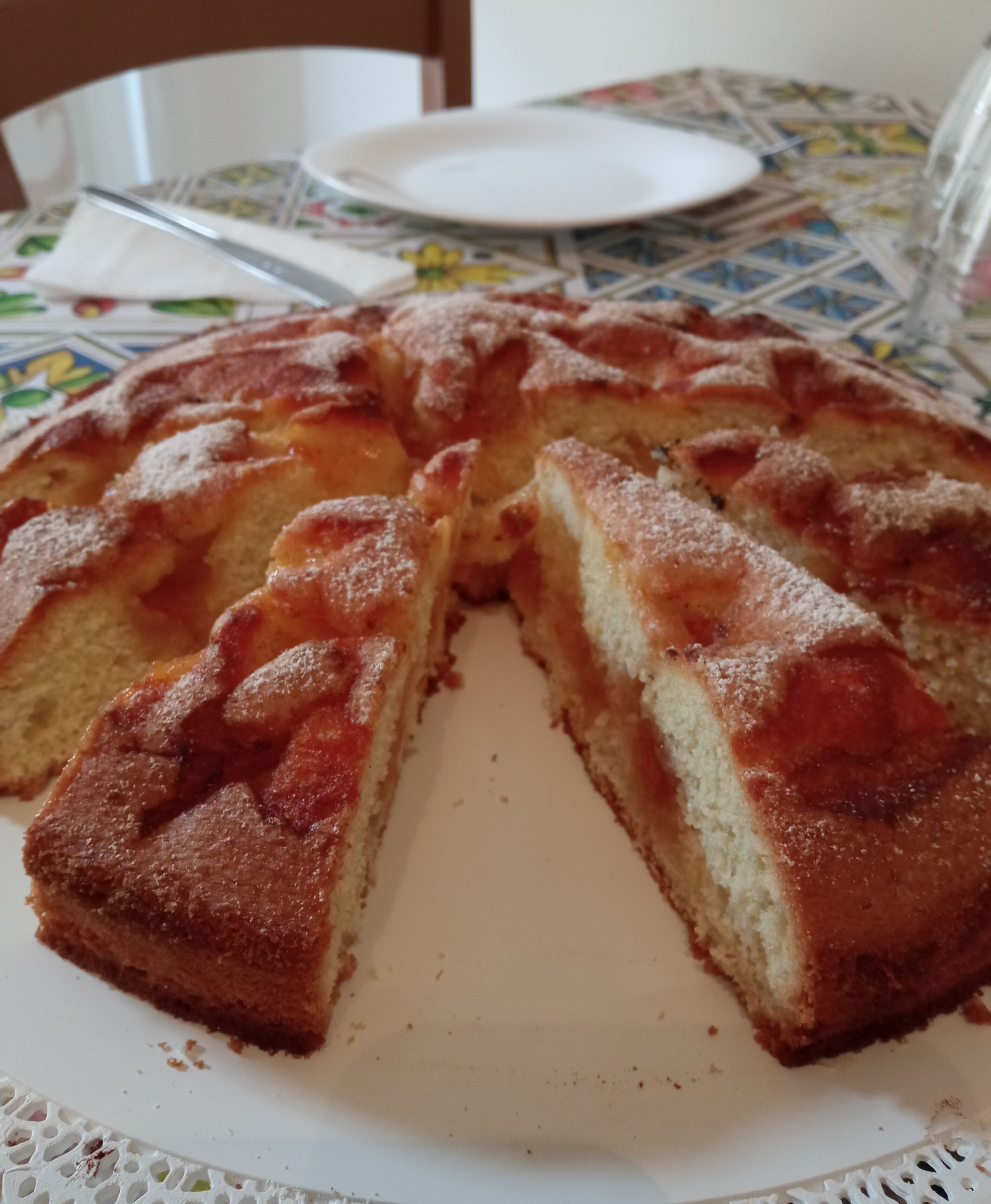Un pastel redondo con una porción faltante, sobre un plato blanco sobre una mesa con mantel estampado.