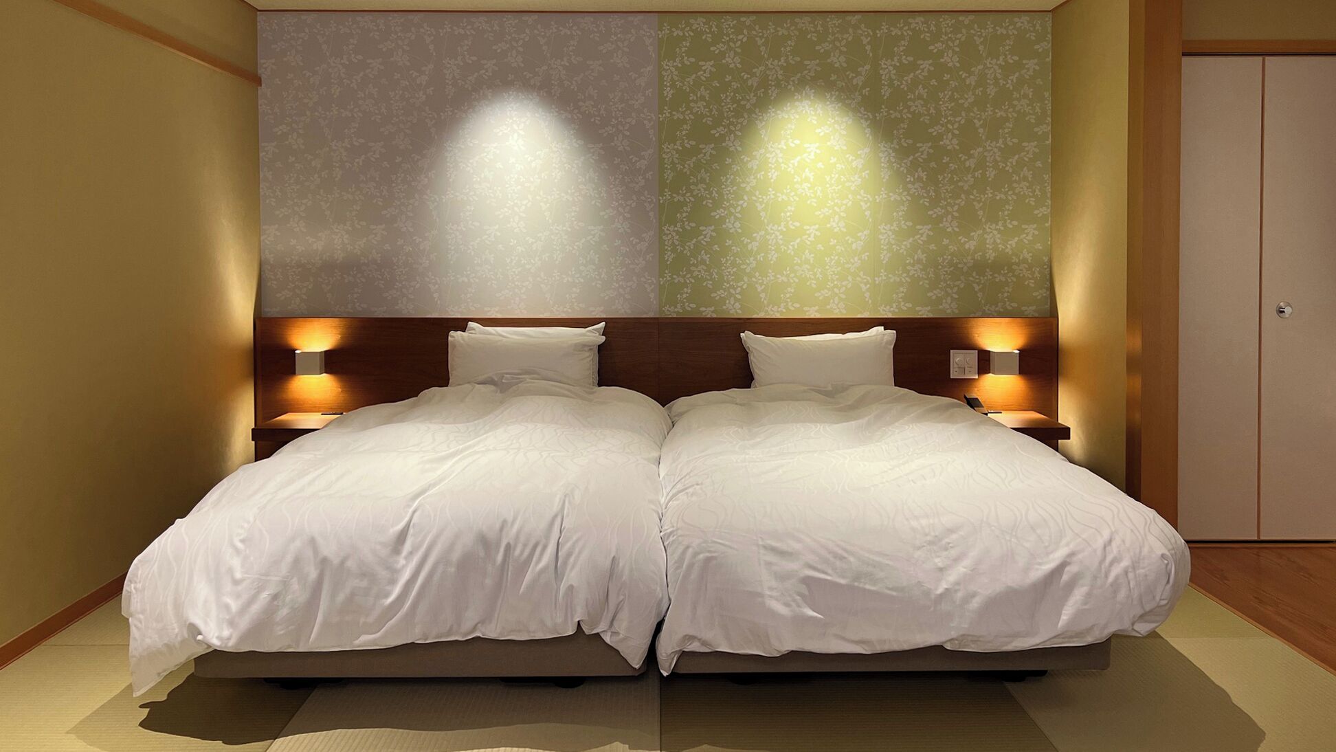 A double bed with two nightstands, each with a wall-mounted lamp, in a room with floral wallpaper and a wooden floor.