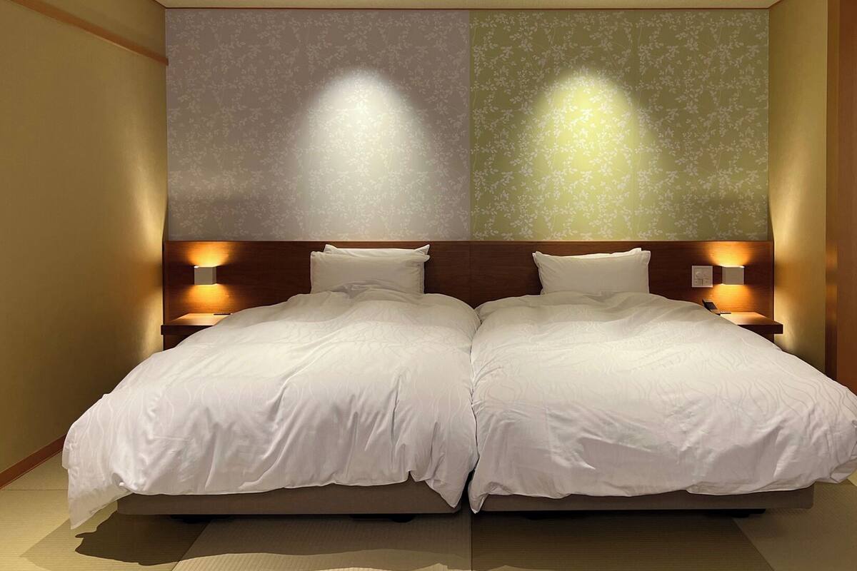 Ein Doppelbett mit zwei Nachttischen, jeweils mit einer Wandlampe, in einem Zimmer mit floralem Wandpapier und Holzboden.