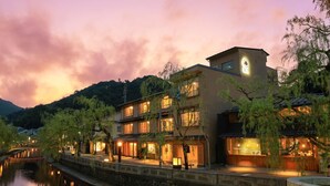 A traditional multi-story building with a river in the foreground, stone walls, and a mountainous backdrop.