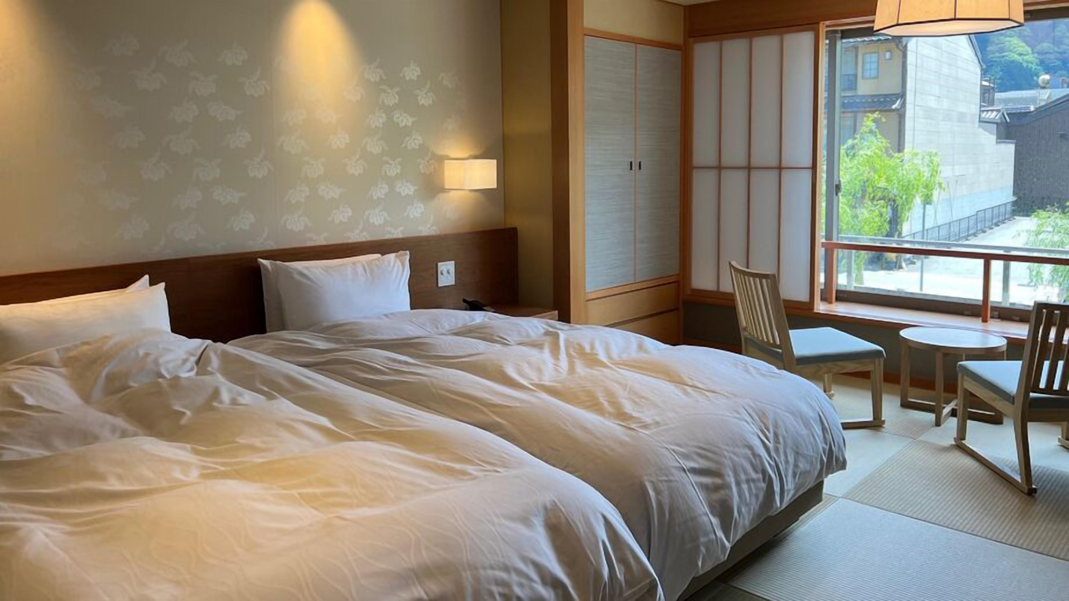 A hotel room with a large bed, a small table, a chair, and a view of buildings through a window.