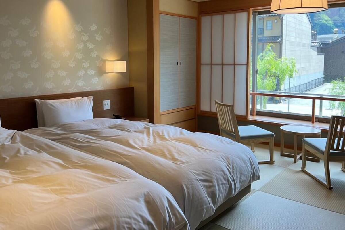 Ein Hotelzimmer mit einem großen Bett, einem kleinen Tisch, einem Stuhl und Blick auf Gebäude durch ein Fenster.