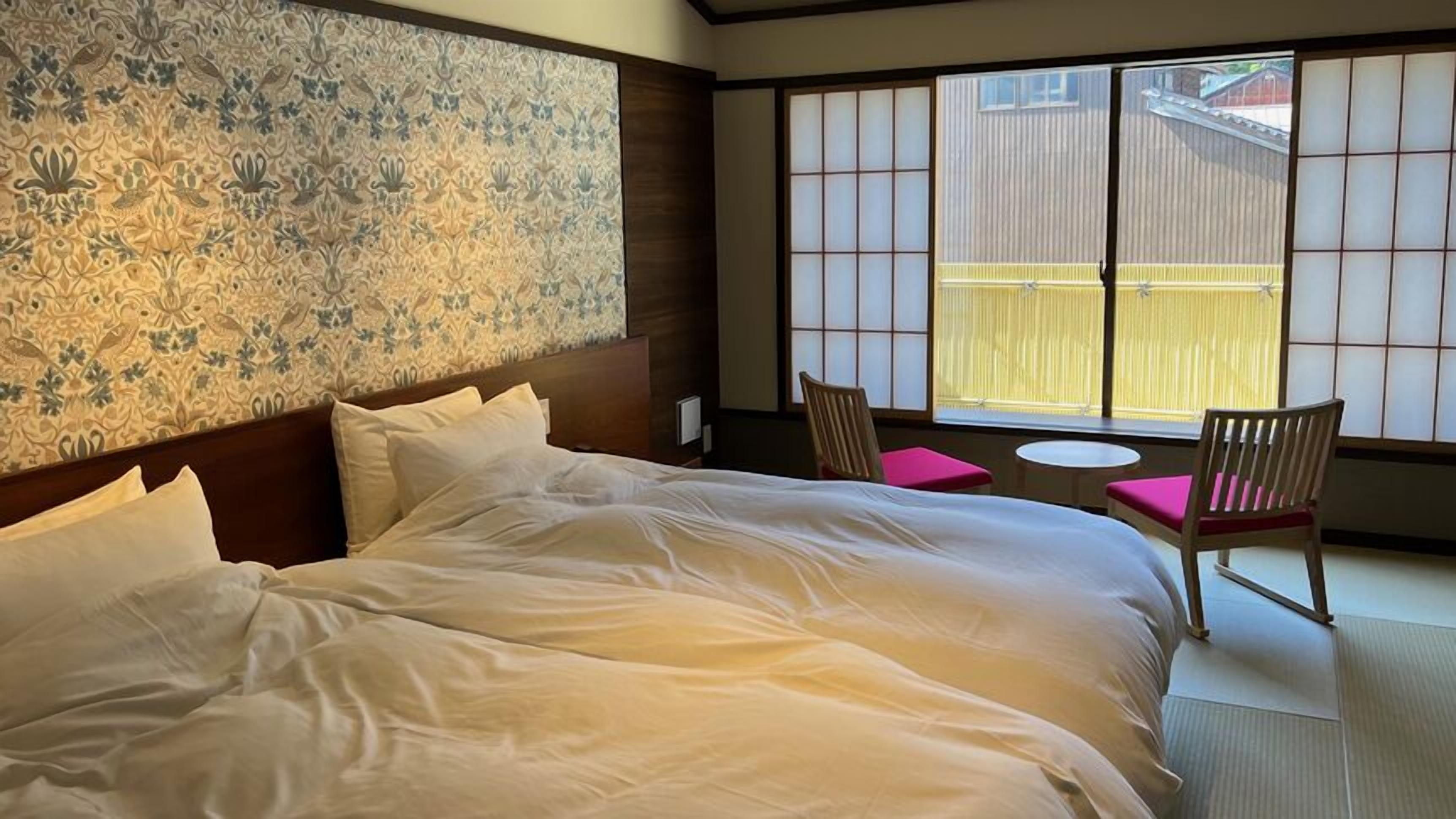 A hotel room with a large bed, two chairs, a small table, and a view through a shoji screen.