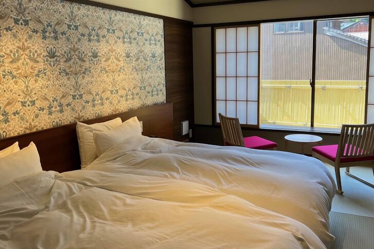 Ein Hotelzimmer mit einem großen Bett, zwei Stühlen, einem kleinen Tisch und Blick durch einen Shoji-Bildschirm.