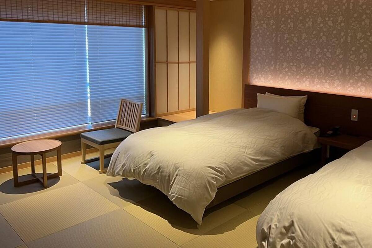 Ein Hotelzimmer mit einem großen Bett, einem Sessel, einem kleinen Tisch und einem Fenster mit Jalousien.