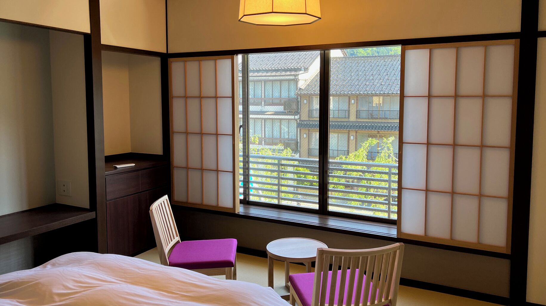 A traditional Japanese room with tatami flooring, a low table, two chairs, a bed, and a view of a traditional building through shoji screens.