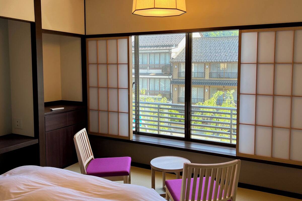 Ein traditioneller japanischer Raum mit Tatami-Böden, einem niedrigen Tisch, zwei Stühlen, einem Bett und Blick auf ein traditionelles Gebäude durch Shoji-Schirme.