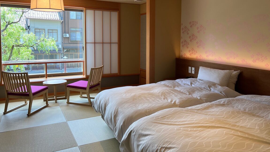 A hotel room with a large bed, a small table, two chairs, a view through a large window, and a traditional Japanese sliding door.