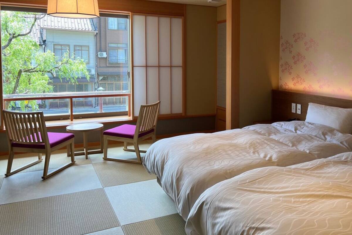 Ein Hotelzimmer mit einem großen Bett, einem kleinen Tisch, zwei Stühlen, Blick durch ein großes Fenster und einer traditionellen japanischen Schiebetür.
