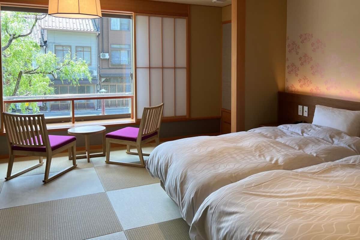 A hotel room with a large bed, a small table, two chairs, a view through a large window, and a traditional Japanese sliding door.