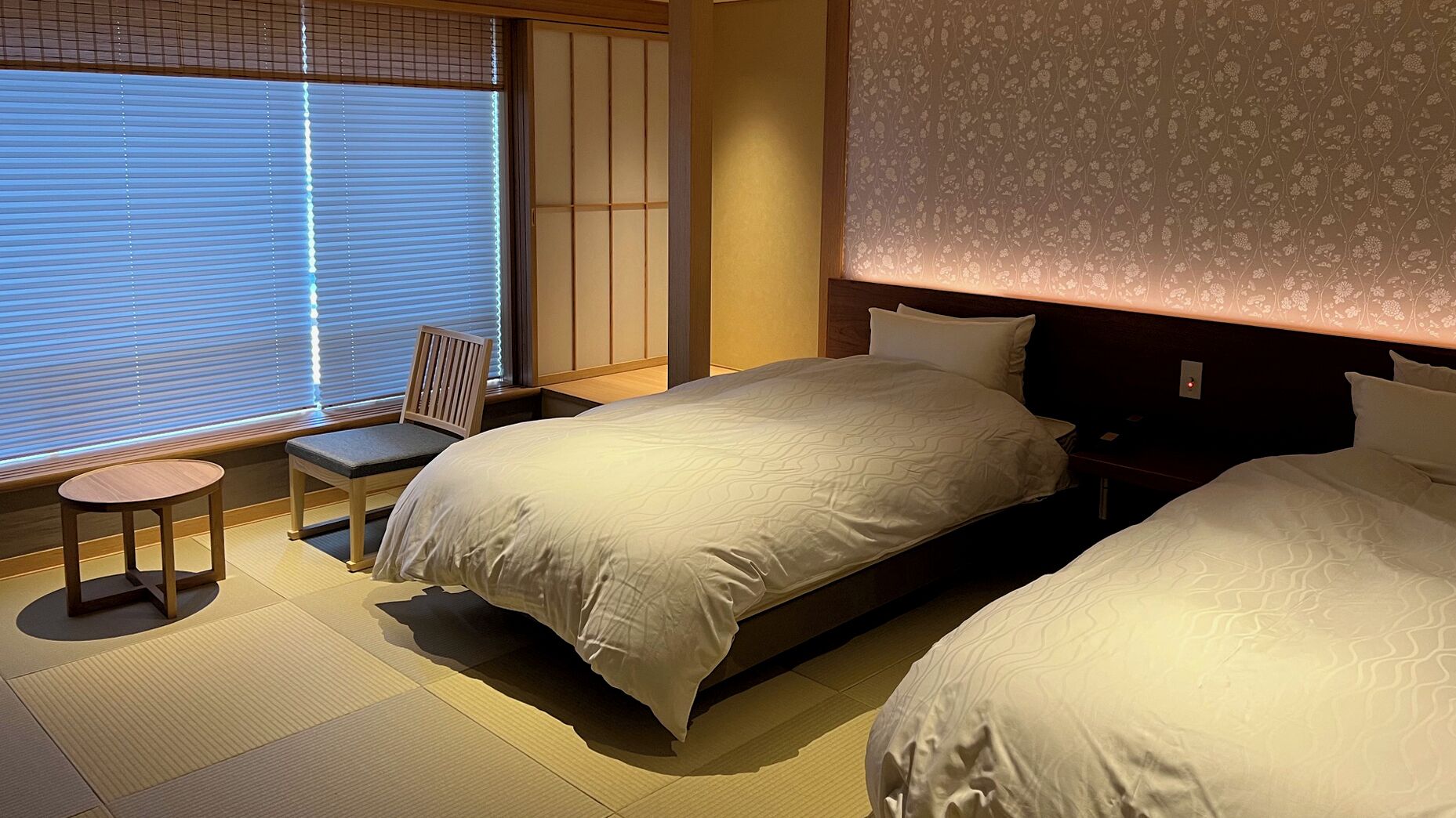 A hotel room with a bed, a chair, a small table, and a window with blinds.