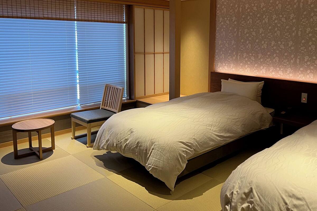 Ein Hotelzimmer mit einem Bett, einem Sessel, einem kleinen Tisch und einem Fenster mit Jalousien.