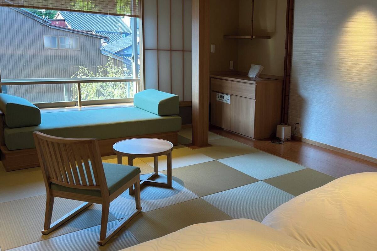 Ein traditioneller japanischer Raum mit Tatami-Bodenbelag, niedrigem Tisch, Sitzbank mit Kissen und einem Bett.