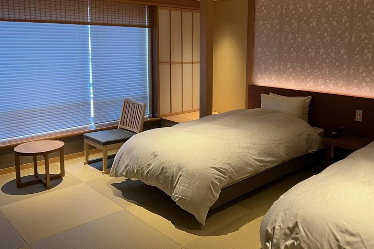 A hotel room with a large bed, a chair, a small table, and a window with blinds.