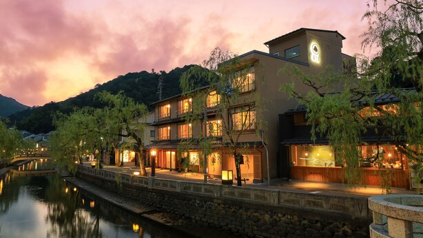 A traditional building with multiple floors, a river in the foreground, and a mountainous backdrop.