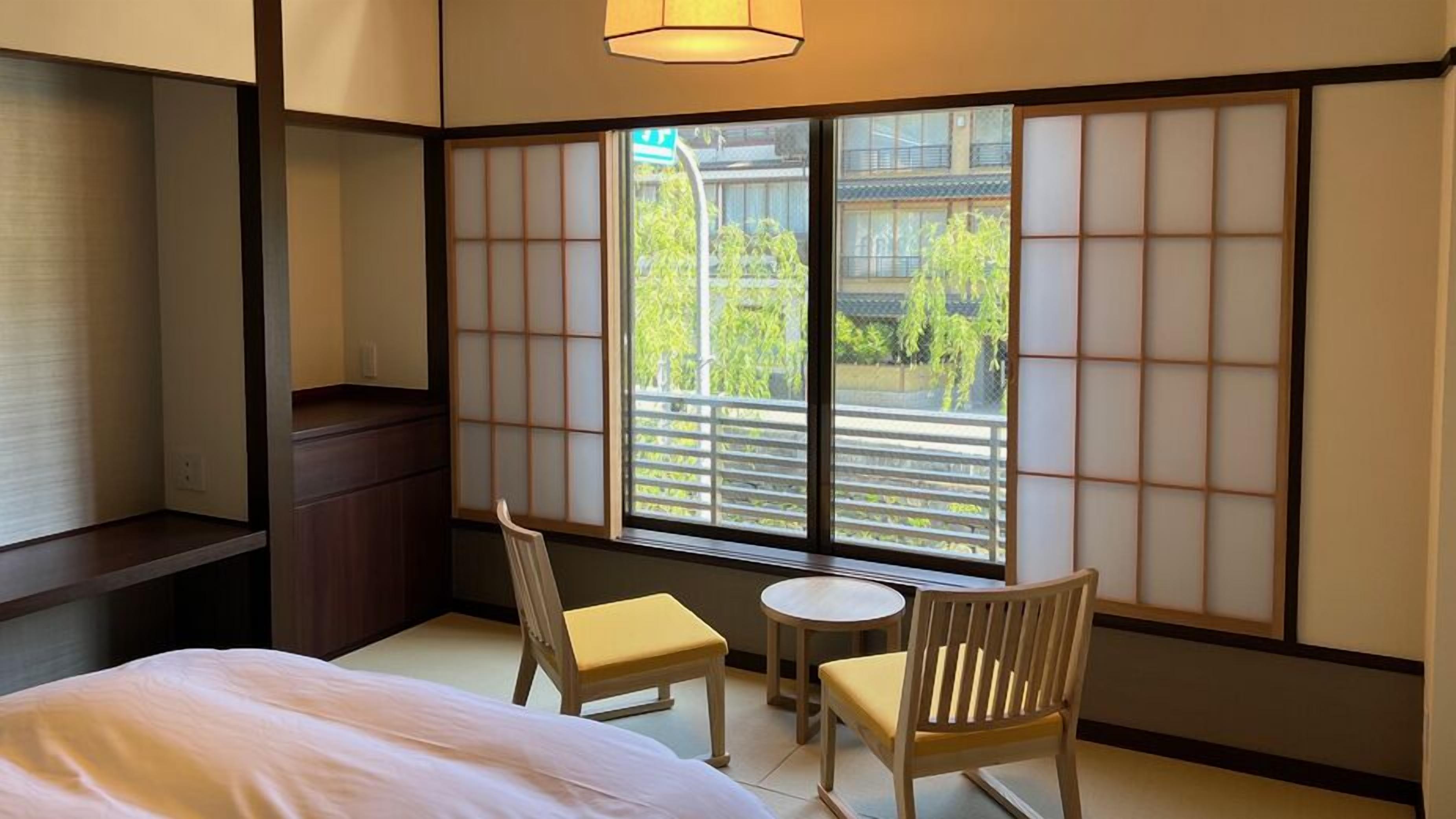 A traditional Japanese-style room with tatami flooring, a low bed, a small round table, two wooden chairs, and a sliding shoji screen.
