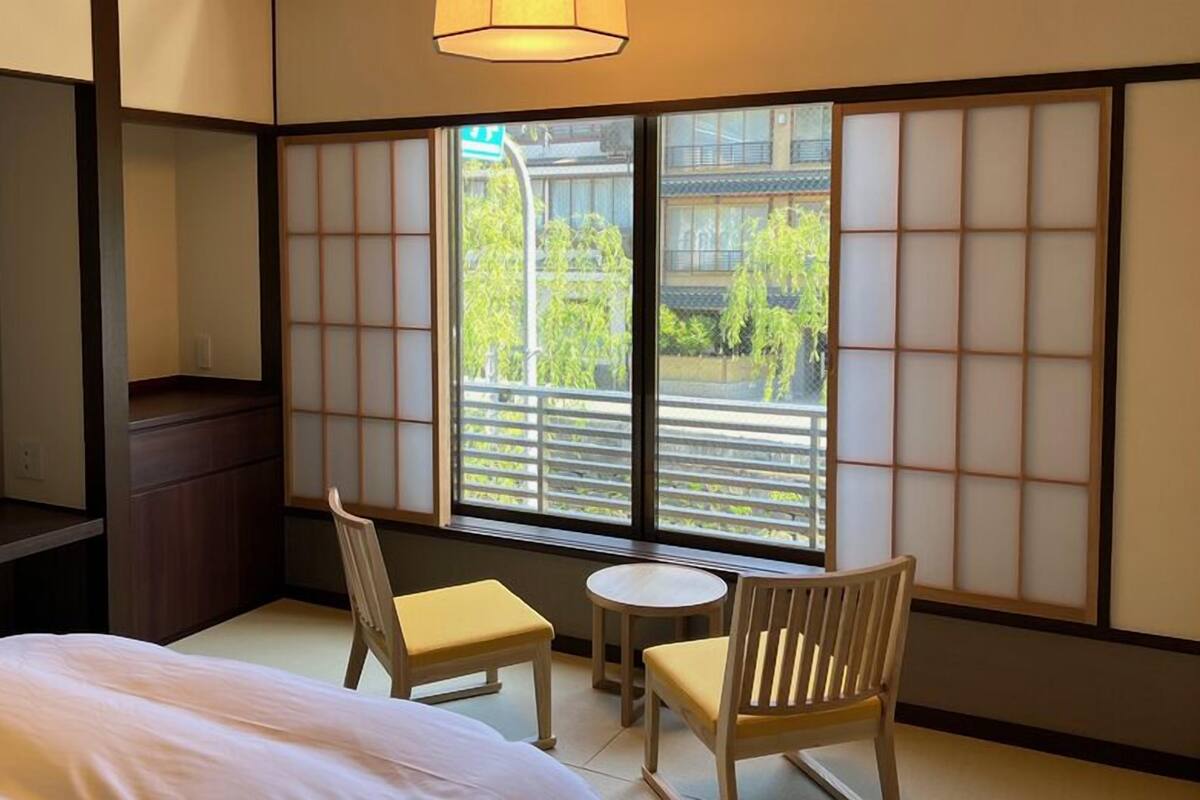Ein traditioneller japanischer Raum mit Tatami-Bodenbelag, einem niedrigen Bett, einem kleinen runden Tisch, zwei Holzsesseln und einer verschiebbaren Shoji-Schirmwand.
