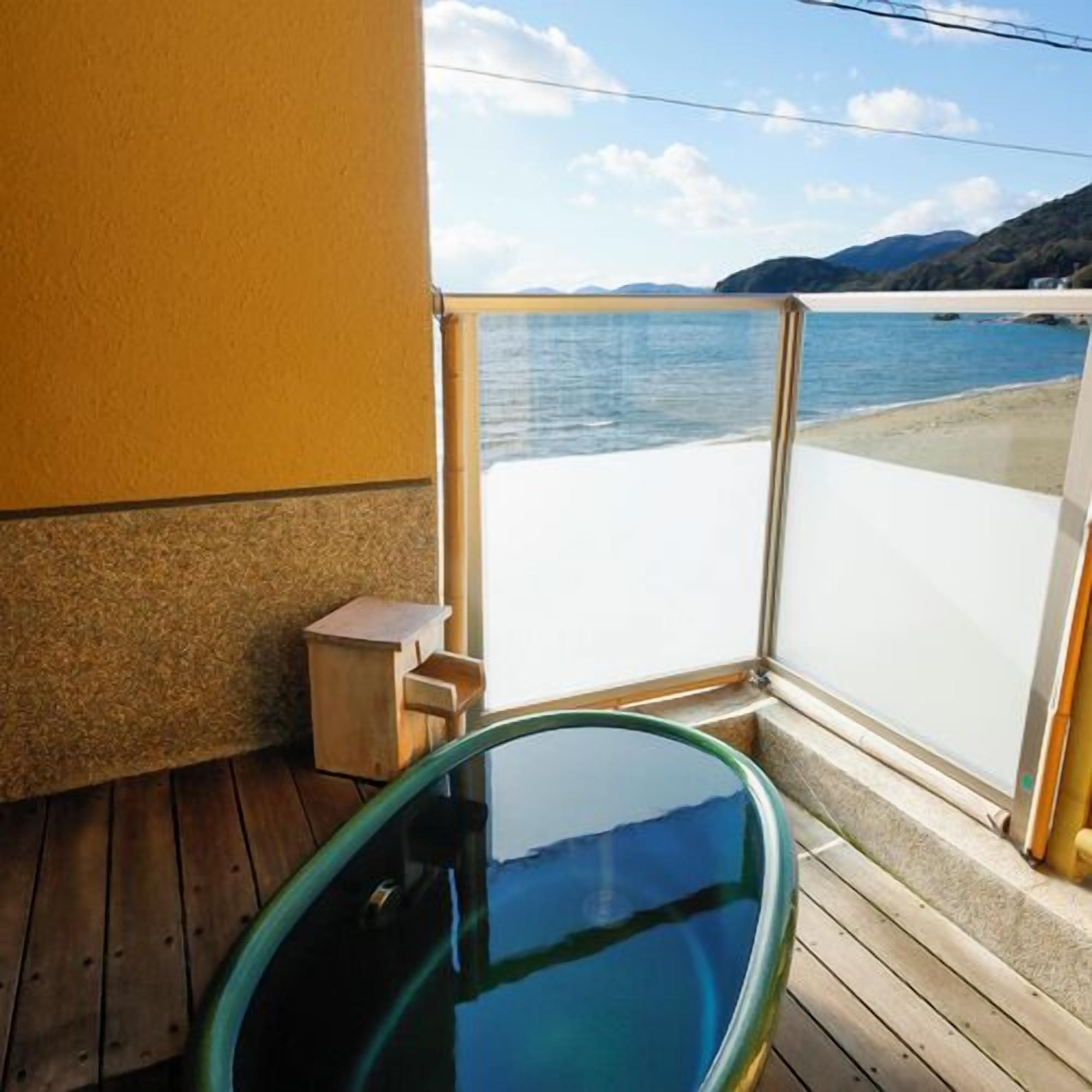 A wooden deck with a glass-enclosed hot tub, a small wooden stool, and a view of the sea and mountains.