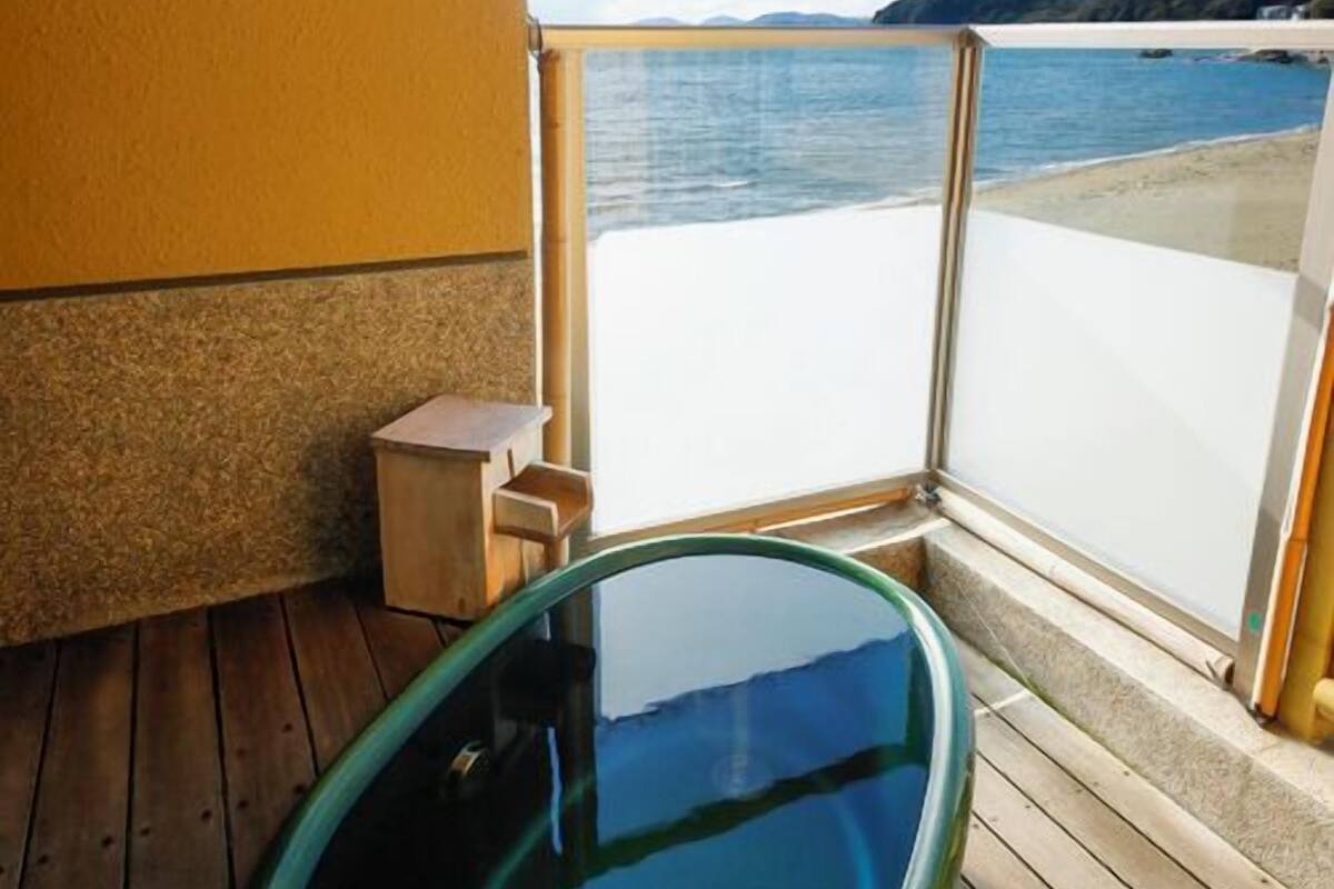 A wooden deck with a glass-enclosed hot tub, a small wooden stool, and a view of the sea and mountains.