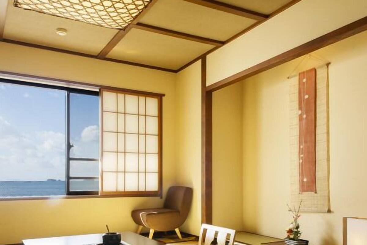 A traditional Japanese room with tatami flooring, a low table with a tea set, a chair, and a window with a view of the sea.