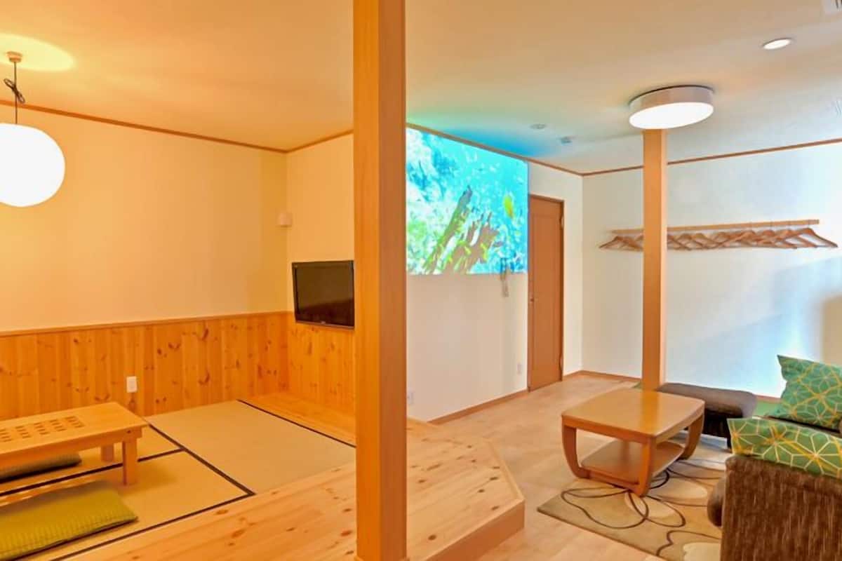 Perinteinen japanilainen huone, jossa on tatamimatto, matala pöytä, tyynyillä varustettu sohva, televisio ja projektorikangas.