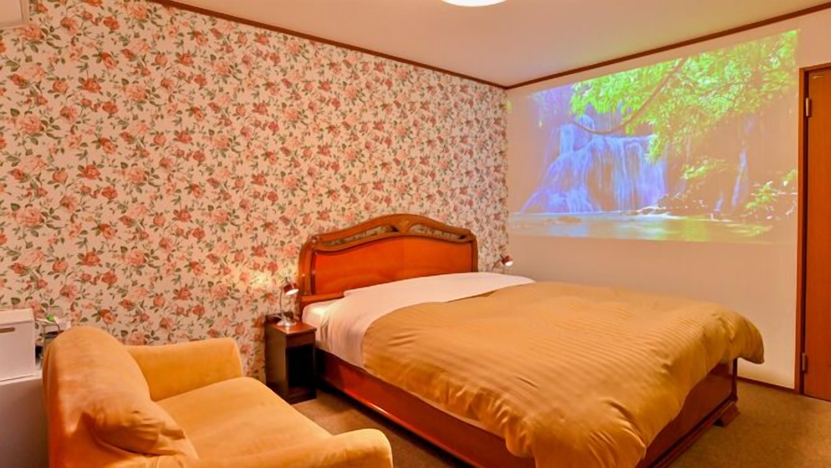 A bedroom with a bed, a chair, a nightstand, and a wall-mounted projector displaying a nature scene.