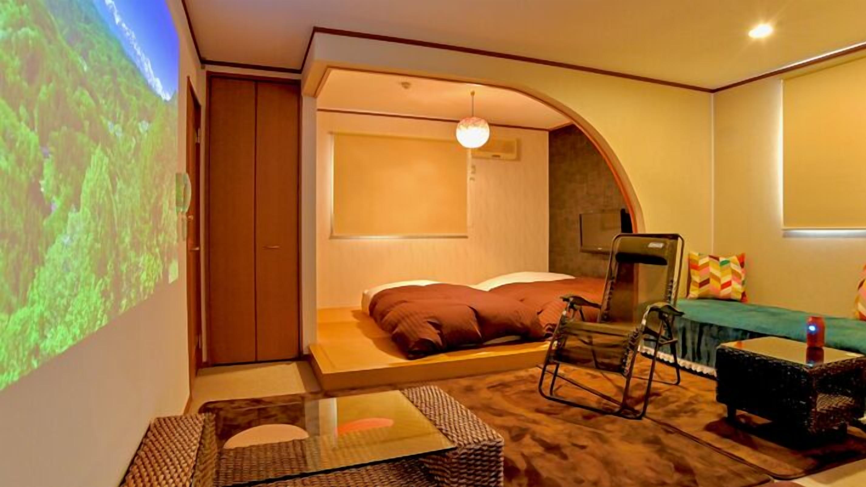 A bedroom with a bed, a chair, a small table, a TV screen, and a wardrobe.