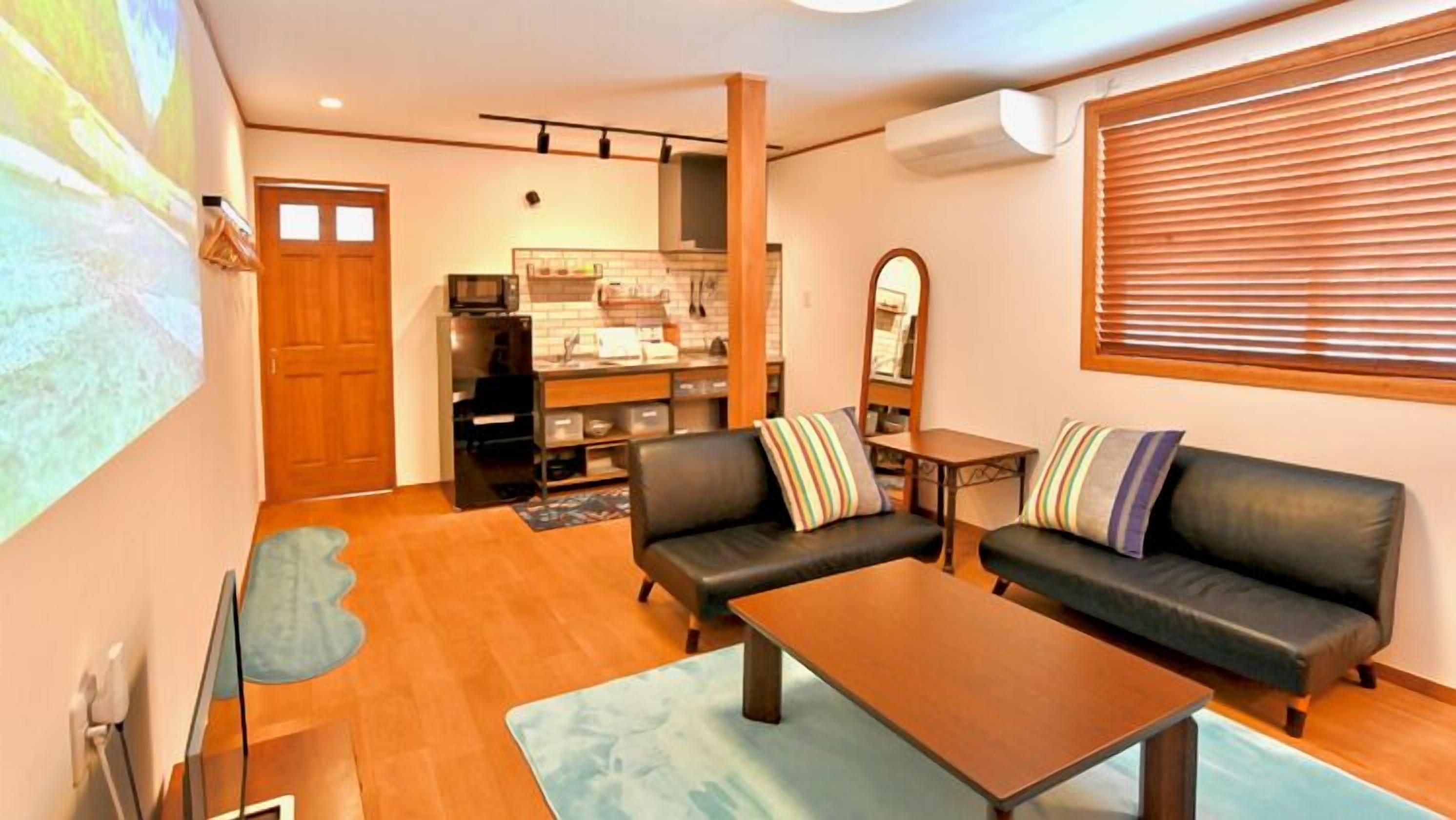 A living room with two sofas, a coffee table, a kitchen area, and a large window with blinds.