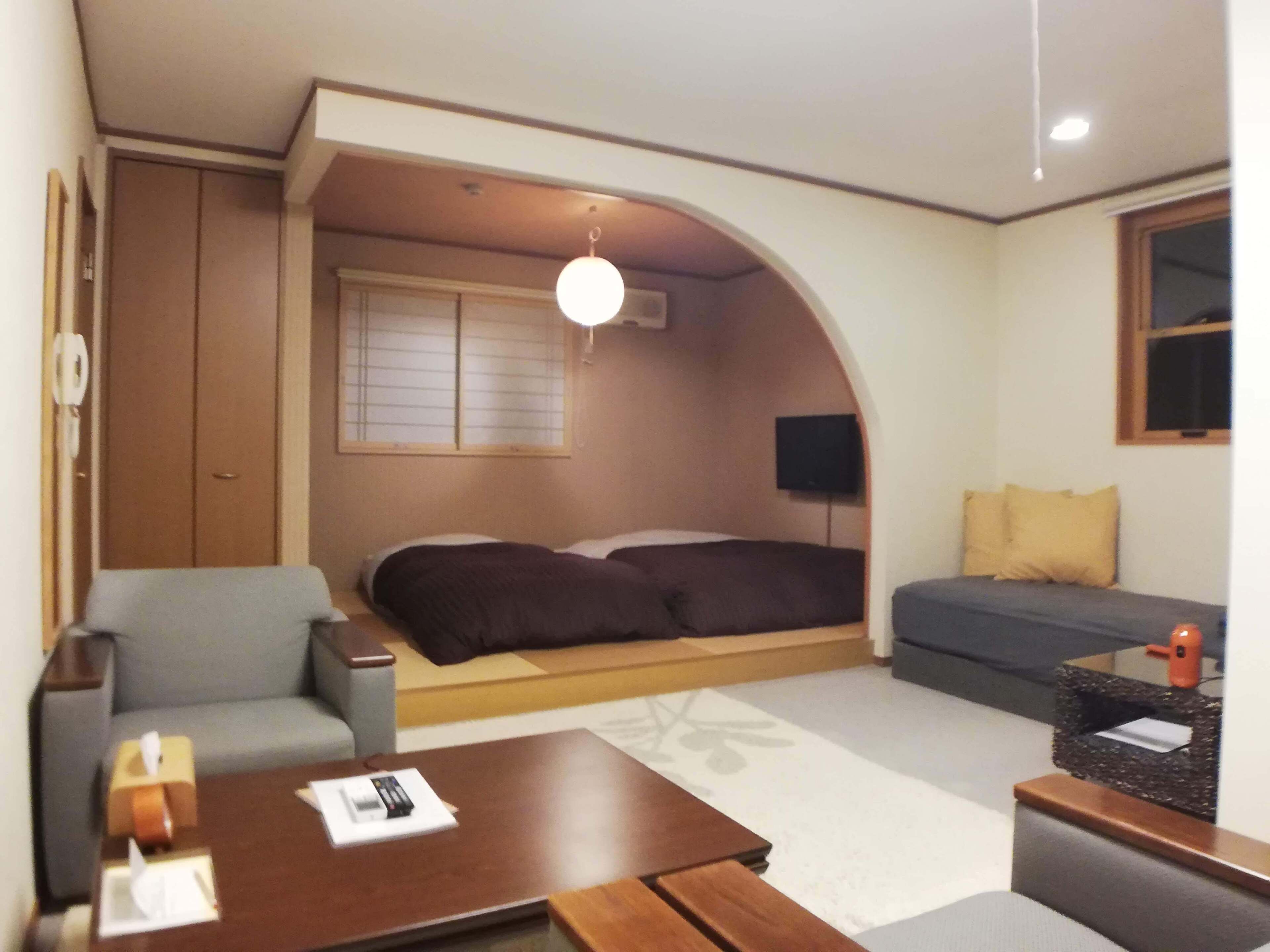 A compact hotel room with a bed, a sofa, a small table, a TV, and a window with blinds.