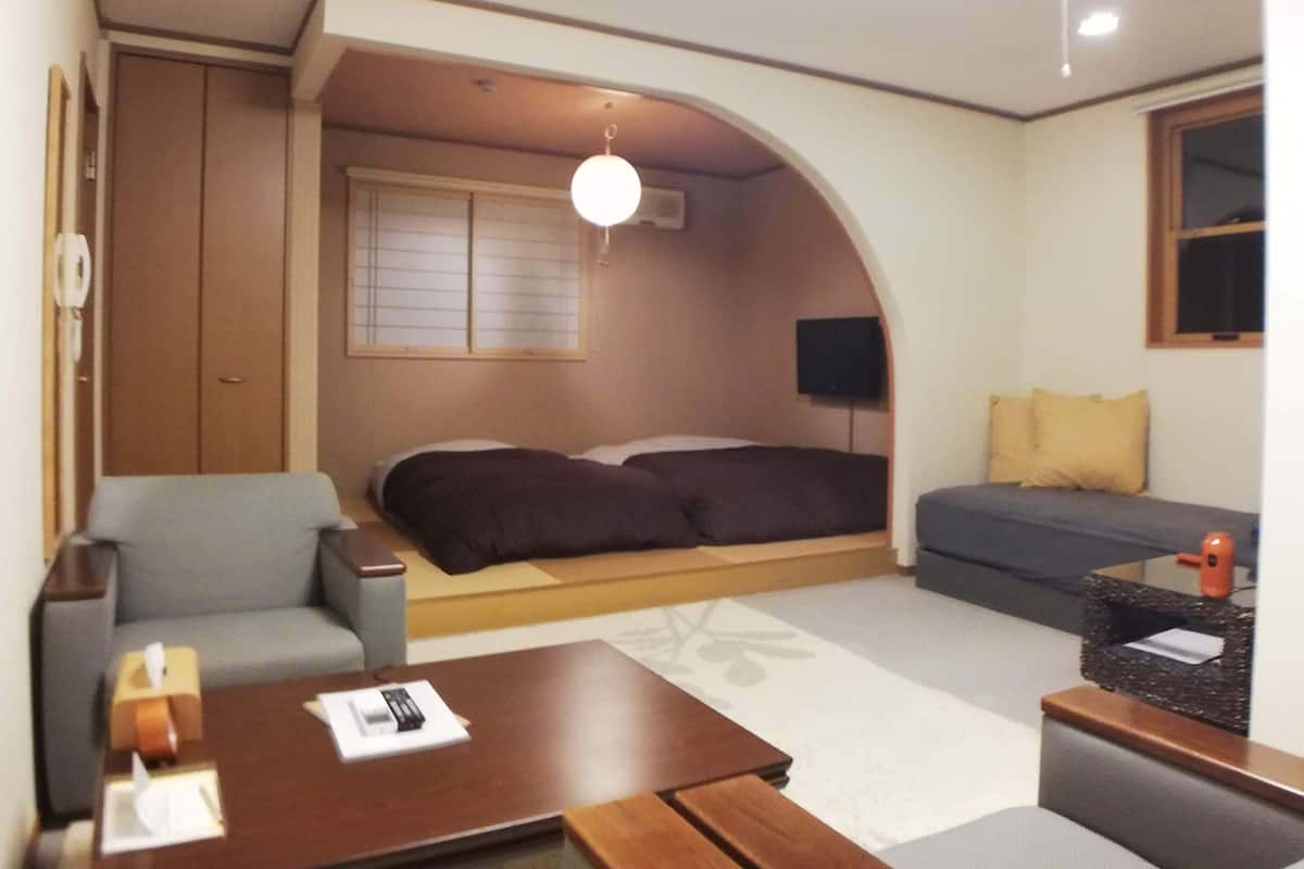 Kompakti hotellihuone, jossa on sänky, sohva, pieni pöytä, televisio ja ikkuna, jossa on sälekaihtimet.