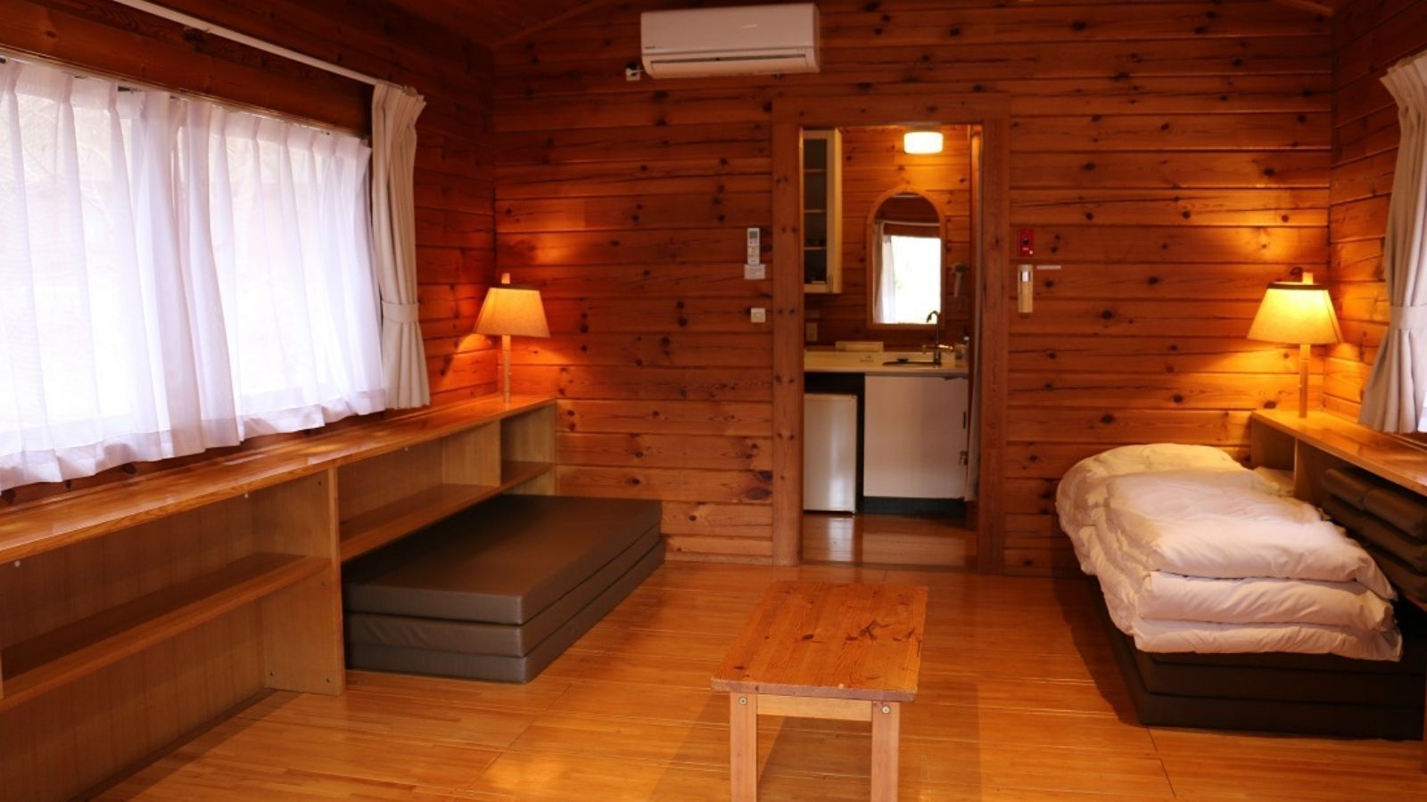 A wooden cabin room with a bed, a small table, a bench, and a kitchenette in the background.