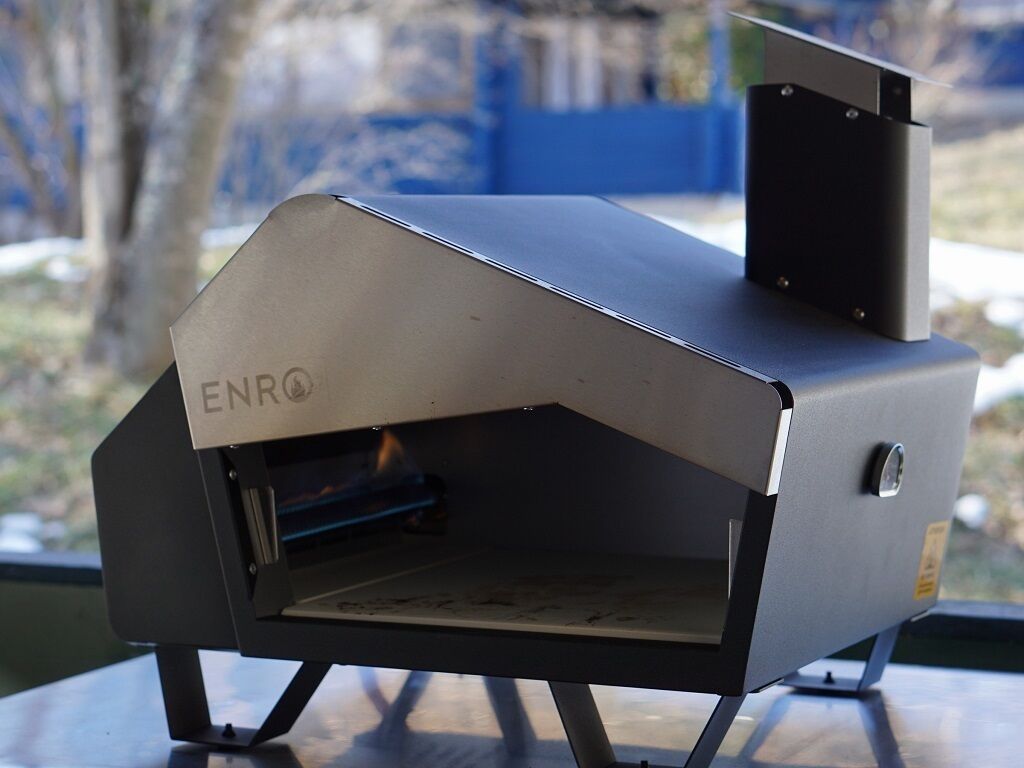 A portable grill with the brand ENRO, placed on a table with a view of trees and a blue wall in the background.