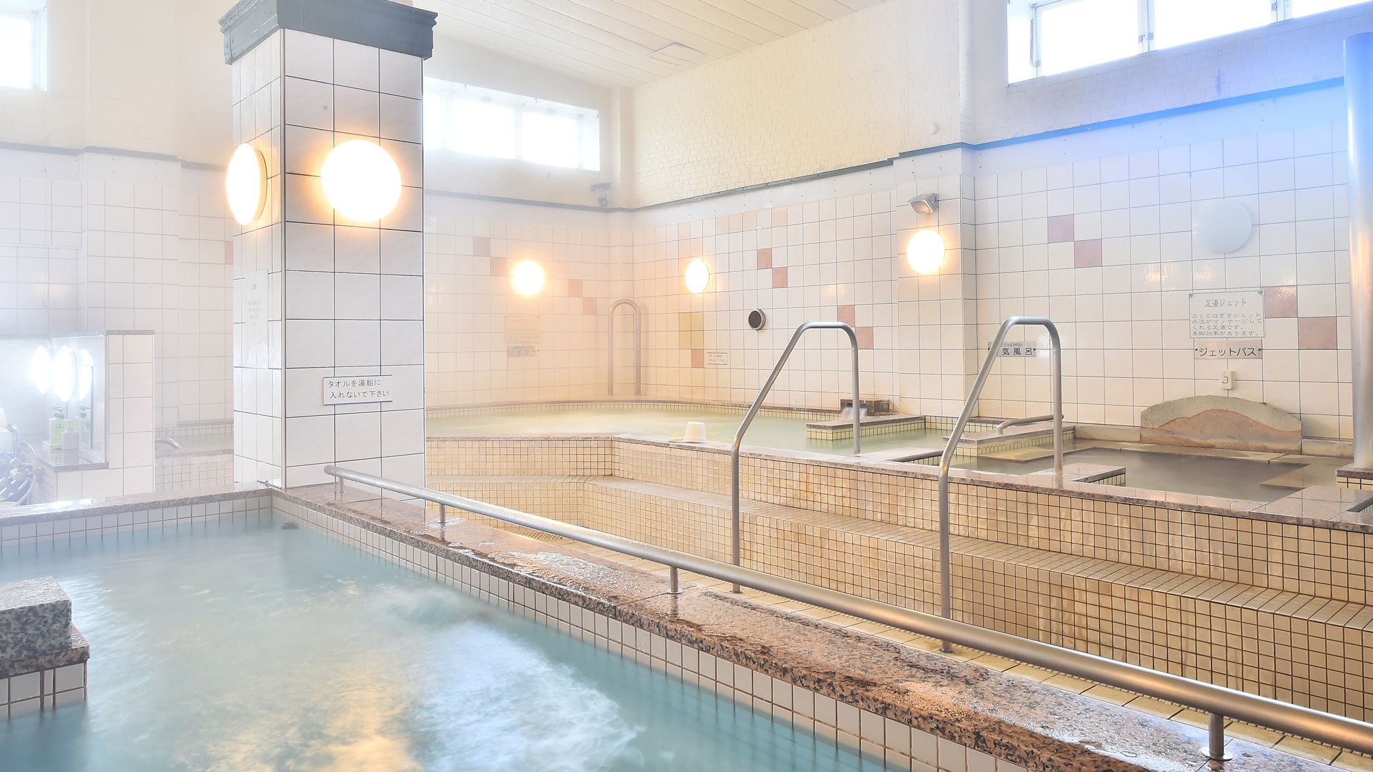 A public bath with multiple hot tubs, tiled walls, and metal railings.